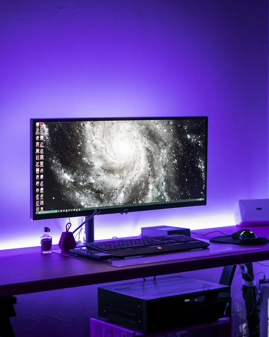 Escritorio con un monitor panorámico que muestra una galaxia, un teclado y un ordenador debajo, iluminado por una luz morada contra la pared