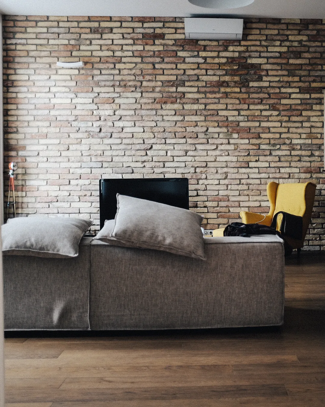 Salón con un sofá gris con cojines delante de una pared de ladrillo visto, un televisor apagado al fondo y un sillón amarillo junto a la ventana