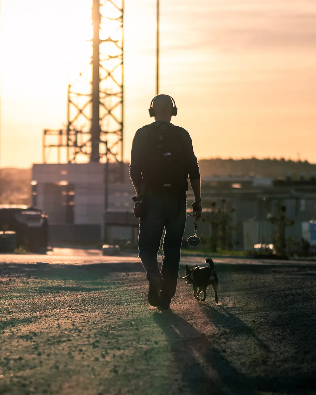 Persona con auriculares paseando a un perro pequeño por una calle al atardecer, a contraluz y con el cielo anaranjado