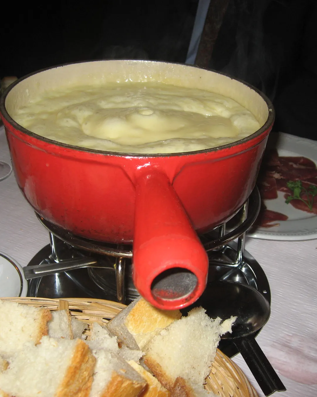 Olla de fondue roja con queso fundido sobre su hornillo en una mesa, con una cesta de pan cortado en dados delante