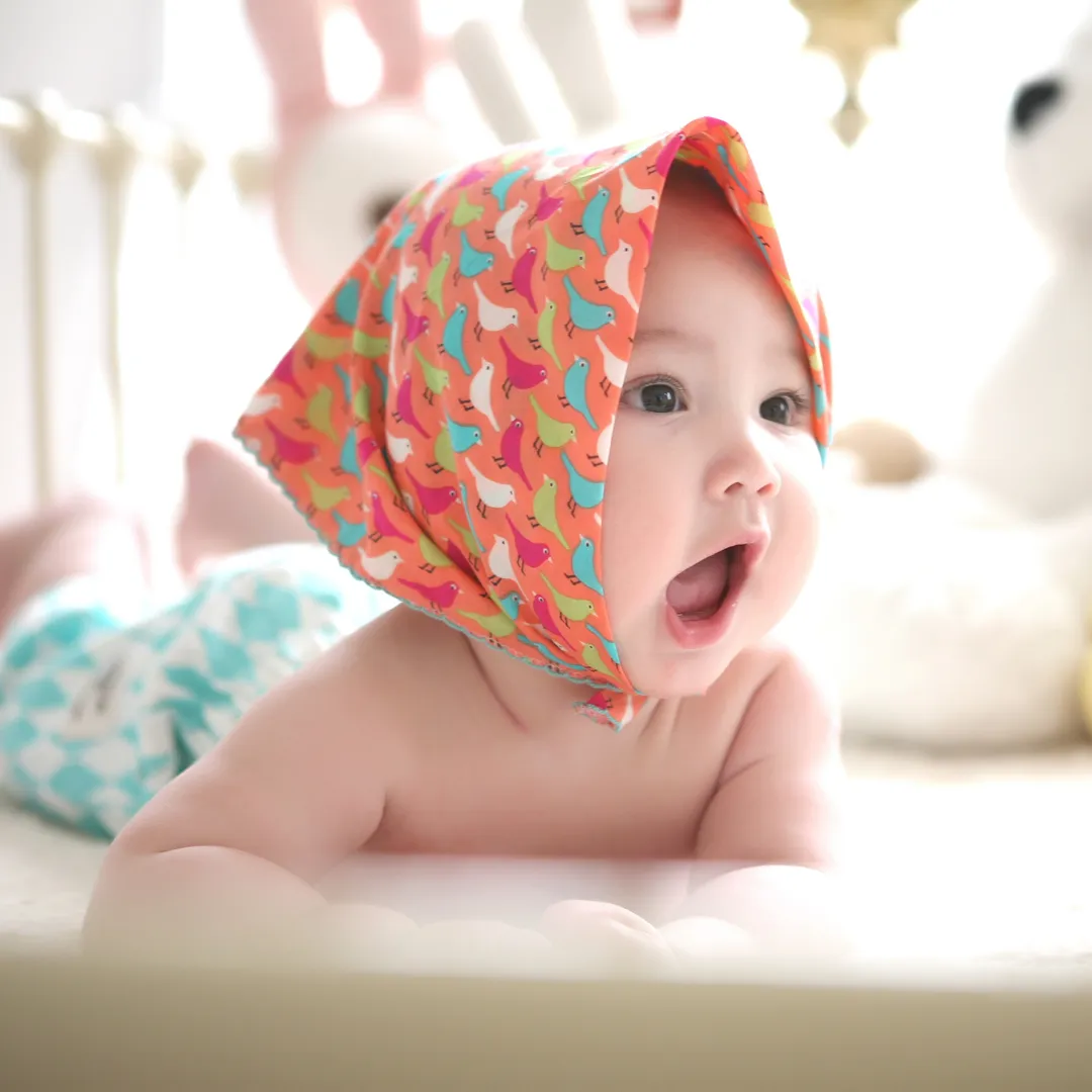 Bebé tumbado boca abajo con un pañuelo de colores en la cabeza y peluches detrás