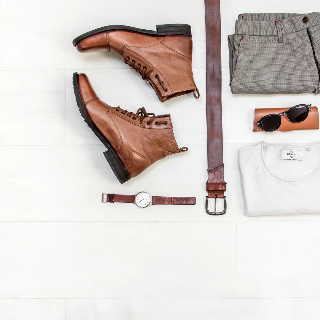 Botas, pantalón, camiseta blanca, cinturón, reloj y gafas de sol ordenados sobre fondo blanco